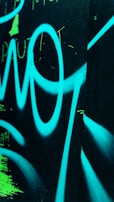Bright blue graffiti or abstract design on a dark surface with visible spray painted letters and shapes, accented with some neon green marks.