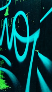 Bright blue graffiti or abstract design on a dark surface with visible spray painted letters and shapes, accented with some neon green marks.