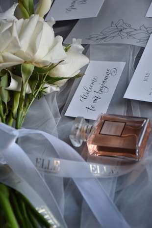 A bouquet of white roses is laid on a soft tulle fabric, accompanied by elegantly styled handwritten cards with phrases like 'Welcome to our beginning'. A bottle of perfume with a pinkish hue is positioned nearby, and a ribbon with the word 'ELLE' is draped gracefully across the scene.