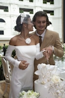 A couple is joyfully cutting a wedding cake together. The bride is wearing an elegant white dress with a floral accessory and a veil, while the groom is dressed in a beige suit with a white shirt and brown tie. They are surrounded by floral decorations and are in an upscale setting with white walls and greenery.