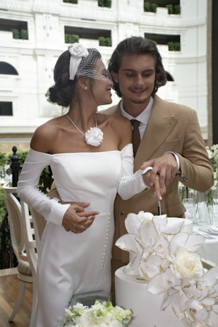 A couple is joyfully cutting a wedding cake together. The bride is wearing an elegant white dress with a floral accessory and a veil, while the groom is dressed in a beige suit with a white shirt and brown tie. They are surrounded by floral decorations and are in an upscale setting with white walls and greenery.