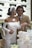A couple is joyfully cutting a wedding cake together. The bride is wearing an elegant white dress with a floral accessory and a veil, while the groom is dressed in a beige suit with a white shirt and brown tie. They are surrounded by floral decorations and are in an upscale setting with white walls and greenery.