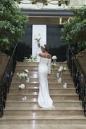 A woman in a white gown stands on a staircase decorated with white flowers and candles. She holds a bouquet in one hand, surrounded by indoor plants and elegant black railings. A decorative panel and a large 'L' are visible in the background.