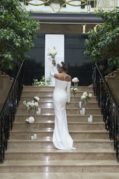 A woman in a white gown stands on a staircase decorated with white flowers and candles. She holds a bouquet in one hand, surrounded by indoor plants and elegant black railings. A decorative panel and a large 'L' are visible in the background.