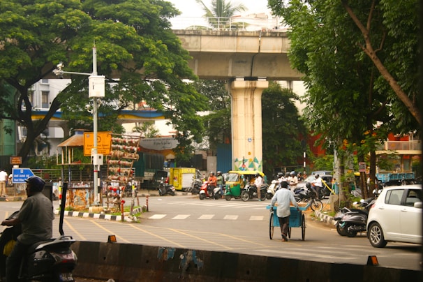 A bustling urban street scene featuring a busy intersection with multiple vehicles, including scooters, motorcycles, and an auto rickshaw. A man is pushing a cart across the road, while greenery from large trees and plants frames the scene. Elevated transportation infrastructure is visible overhead, and various road signs and traffic signals are present.