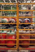 Several gas cylinders are secured in the back of a truck with yellow metal railings. The cylinders are primarily red and green, with visible brand markings and regulatory information. The vehicle is parked, and vegetation can be seen in the background.