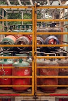 Several gas cylinders are secured in the back of a truck with yellow metal railings. The cylinders are primarily red and green, with visible brand markings and regulatory information. The vehicle is parked, and vegetation can be seen in the background.