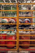Several gas cylinders are secured in the back of a truck with yellow metal railings. The cylinders are primarily red and green, with visible brand markings and regulatory information. The vehicle is parked, and vegetation can be seen in the background.