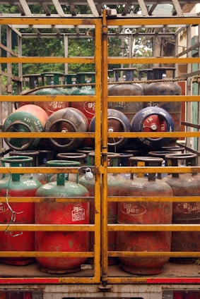 Several gas cylinders are secured in the back of a truck with yellow metal railings. The cylinders are primarily red and green, with visible brand markings and regulatory information. The vehicle is parked, and vegetation can be seen in the background.