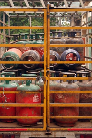 Several gas cylinders are secured in the back of a truck with yellow metal railings. The cylinders are primarily red and green, with visible brand markings and regulatory information. The vehicle is parked, and vegetation can be seen in the background.