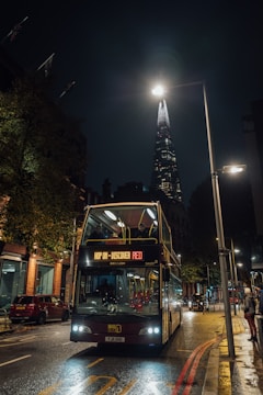A double-decker bus travels along a dimly lit city street at night, with the iconic Shard skyscraper illuminated in the background. The bus displays a sign 'HOP ON - DISCOVER RED'. Trees and buildings line the street, while several cars and pedestrians are present. Streetlights create a warm ambiance, reflecting on the wet pavement.