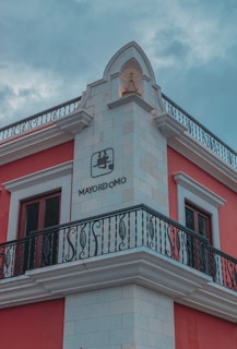 A corner view of a building with red walls and white accents, including ornate architectural details at the top. There is a black wrought iron balcony and rectangular windows with white frames. The word 'MAYORDOMO' is clearly visible along with a logo. The sky is overcast, giving a somewhat moody ambiance to the scene.