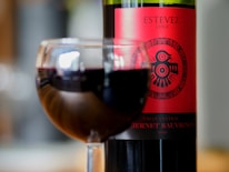 A glass filled with red wine is in the foreground, with a bottle of Cabernet Sauvignon wine set in the background. The bottle label is red with decorative black symbols, prominently displaying the name Estevez and indicating it is from Chile.