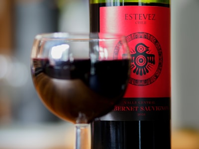 A glass filled with red wine is in the foreground, with a bottle of Cabernet Sauvignon wine set in the background. The bottle label is red with decorative black symbols, prominently displaying the name Estevez and indicating it is from Chile.