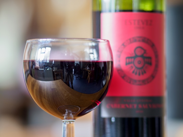 A close-up of a glass filled with red wine, with a bottle of Cabernet Sauvignon positioned in the background. The wine glass reflects the interior of a room, showcasing details like furniture and windows. The wine bottle has a prominent red label featuring decorative graphics and text.