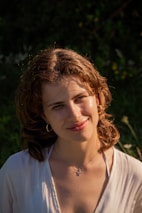 Elegant woman wearing floral earrings, smiling gently in natural sunlight outdoors.