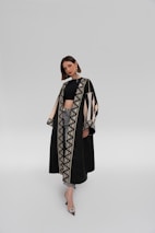 A woman stands confidently wearing a long, elegant black coat with intricate geometric patterns along the edges. The coat is accented with beige and black designs, and she's paired it with a black top and jeans. She sports large, stylish earrings and pointed heels, against a plain, light grey background.