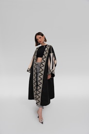 A woman stands confidently wearing a long, elegant black coat with intricate geometric patterns along the edges. The coat is accented with beige and black designs, and she's paired it with a black top and jeans. She sports large, stylish earrings and pointed heels, against a plain, light grey background.
