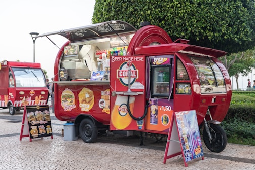 A vibrant red food truck with an eye-catching design, parked on a cobblestone street. The truck displays various food advertisements including burgers and cheese rolls. There are also drink and snack options visible. The truck is adorned with bright signage advertising beverages for 1.50 euros. A menu board on an easel is placed beside the truck, showcasing different food items. Another similar food truck is parked in the background. The scene is set in an outdoor area with a green, leafy tree nearby.