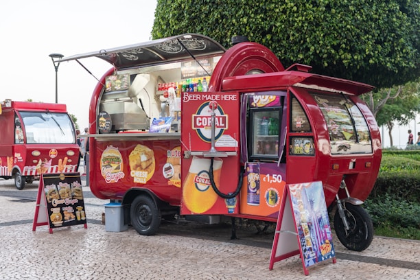 A vibrant red food truck with an eye-catching design, parked on a cobblestone street. The truck displays various food advertisements including burgers and cheese rolls. There are also drink and snack options visible. The truck is adorned with bright signage advertising beverages for 1.50 euros. A menu board on an easel is placed beside the truck, showcasing different food items. Another similar food truck is parked in the background. The scene is set in an outdoor area with a green, leafy tree nearby.