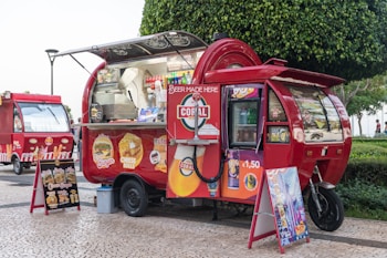 A vibrant red food truck with an eye-catching design, parked on a cobblestone street. The truck displays various food advertisements including burgers and cheese rolls. There are also drink and snack options visible. The truck is adorned with bright signage advertising beverages for 1.50 euros. A menu board on an easel is placed beside the truck, showcasing different food items. Another similar food truck is parked in the background. The scene is set in an outdoor area with a green, leafy tree nearby.