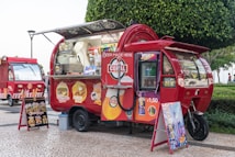 A vibrant red food truck with an eye-catching design, parked on a cobblestone street. The truck displays various food advertisements including burgers and cheese rolls. There are also drink and snack options visible. The truck is adorned with bright signage advertising beverages for 1.50 euros. A menu board on an easel is placed beside the truck, showcasing different food items. Another similar food truck is parked in the background. The scene is set in an outdoor area with a green, leafy tree nearby.
