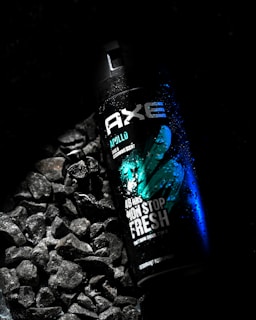 A black can of Axe body spray, labeled 'Apollo,' is placed on a bed of dark wet stones. The can features green and blue accents with water droplets on its surface, creating a fresh and invigorating feel.