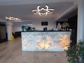 A modern reception area with a marble desk illuminated from below. The ceiling features unique, abstract light fixtures that resemble cloud shapes. There is a person behind the desk, seemingly working on a computer. To the right of the desk are some decorative items, including a plant, and a rack with informational flyers or brochures. There is a caution sign on the left side of the image and comfortable seating is visible in the background.