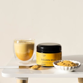 A glass cup filled with a frothy, golden beverage sits on a white table next to a jar labeled 'Golden Milk'. A small, ridged white dish contains a mound of powder, and a golden spoon rests beside it, also filled with the same powder. The overall setting is minimalistic and well-lit.
