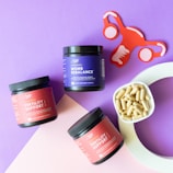 The image contains three jars of dietary supplements labeled 'Womb Rebalance' and 'Fertility Support,' placed against a purple background. A small white bowl filled with capsules is nearby. There is also a red and white paper cutout of a uterus, adding a thematic element to the composition.