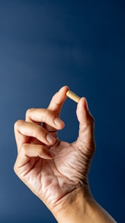 A hand with fingers gracefully holding a small capsule against a solid dark blue background. The skin's texture is visible, highlighting a sense of realism and focus.