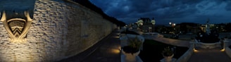A nighttime scene of a luxurious estate with a stone wall featuring an ornate relief. The walkway is adorned with decorative plants in stone planters, leading to a grand building with turreted roofs and illuminated windows. Several street lamps light the pathway, creating a warm ambiance. The sky is dark, filled with clouds, suggesting a moody atmosphere.