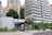 Modern high-rise apartment buildings with a lush green environment surrounding the base. A small kiosk is present on the sidewalk, displaying advertisements and magazines. Tall trees are growing alongside planted shrubs and decorative plants.