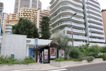 Modern high-rise apartment buildings with a lush green environment surrounding the base. A small kiosk is present on the sidewalk, displaying advertisements and magazines. Tall trees are growing alongside planted shrubs and decorative plants.
