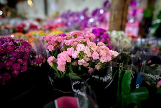 A colorful assortment of flowers arranged in bouquets. The foreground features vibrant pink flowers, while the background is filled with flowers in shades of purple, red, and white. The angle suggests a market or shop display, with a soft focus on the surrounding area.
