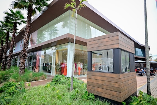 A modern retail store with large glass windows showcasing mannequins dressed in colorful clothing. Palm trees line the sidewalk, and there is a wooden structure in the foreground, possibly a security booth. People are seated in an outdoor dining area to the right.