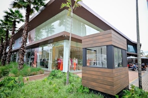 A modern retail store with large glass windows showcasing mannequins dressed in colorful clothing. Palm trees line the sidewalk, and there is a wooden structure in the foreground, possibly a security booth. People are seated in an outdoor dining area to the right.