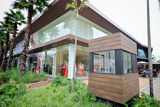 A modern retail store with large glass windows showcasing mannequins dressed in colorful clothing. Palm trees line the sidewalk, and there is a wooden structure in the foreground, possibly a security booth. People are seated in an outdoor dining area to the right.