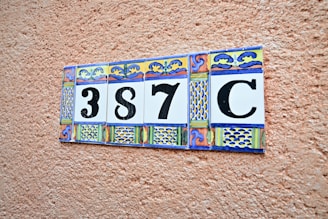 A decorative ceramic tile display with a number '387C' set against a textured terracotta-colored wall. Each tile features ornate patterns with blue, yellow, and red colors.