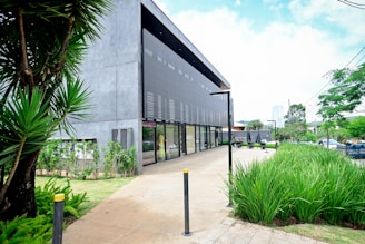 A modern, minimalist building with a concrete facade and large glass windows is surrounded by lush greenery, including tall plants and neatly trimmed grass. The pathway along the building is wide, and there are street lamps and bollards present. The sky is bright, suggesting a sunny day.
