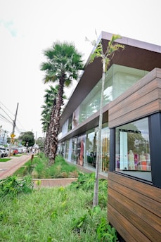 A modern building featuring large glass windows and a wooden facade, surrounded by a lush green landscape with tall palm trees and neatly maintained grass. A busy street runs parallel to the building with numerous cars and some people walking along the sidewalk.