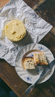 On a wooden table, there is a round piece of butter on crumpled foil, next to a vintage plate with blue floral patterns. The plate holds partially toasted pieces of bread. A knife lies on the plate, facing outward.