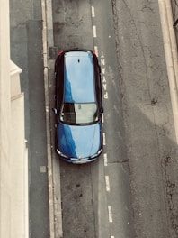 A blue car is parked on the side of a street, seen from an overhead view. The pavement is labeled with the word 'PAYANT' indicating a paid parking area. The surrounding area shows weathered road surfaces with visible cracks and faded lines.