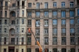 An ornate multi-story building with numerous windows and decorative stone carvings on the facade. A person in orange work attire operates a lift to perform maintenance or cleaning work on the building's exterior.