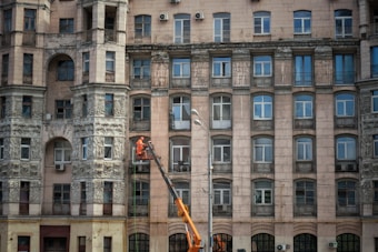 An ornate multi-story building with numerous windows and decorative stone carvings on the facade. A person in orange work attire operates a lift to perform maintenance or cleaning work on the building's exterior.