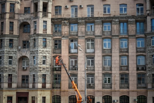An ornate multi-story building with numerous windows and decorative stone carvings on the facade. A person in orange work attire operates a lift to perform maintenance or cleaning work on the building's exterior.