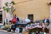 An outdoor market stall displays an assortment of colorful ceramics, bags, and various items. The setting is along a rustic wall with a small window barred with wrought iron. A fig tree leans against the wall, adding greenery to the scene. Items are organized on tables and hanging displays, including bowls, mugs, and fabrics with vibrant patterns.