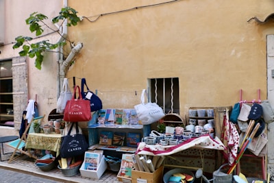 An outdoor market stall displays an assortment of colorful ceramics, bags, and various items. The setting is along a rustic wall with a small window barred with wrought iron. A fig tree leans against the wall, adding greenery to the scene. Items are organized on tables and hanging displays, including bowls, mugs, and fabrics with vibrant patterns.
