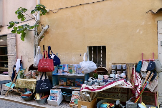 An outdoor market stall displays an assortment of colorful ceramics, bags, and various items. The setting is along a rustic wall with a small window barred with wrought iron. A fig tree leans against the wall, adding greenery to the scene. Items are organized on tables and hanging displays, including bowls, mugs, and fabrics with vibrant patterns.