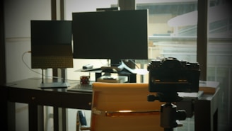 A workspace with two computer screens, a laptop, a camera on a tripod, and a chair. The setting is in front of a large window, allowing natural light to fill the room.